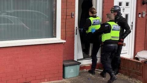 An officer in riot gear stands to the side, a female officer enters a house, a man behind her. It is a red brick house with a white front door and closed blinds.