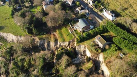 A drone image of a landslide on the Isle of Wight. The birdseye view shows a section of cliff, covered in grass has fallen down, with trees falling down the edge as well as what looks like a red shed. There is a house right on the edge of the cliff and the whole area is lined by trees.