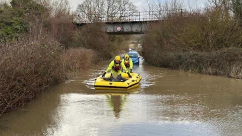 Road submerged in flood water - a man and firefighters are in a yellow inflatable boat with another firefighter guiding it through the water a blue car and bridge are behind them.