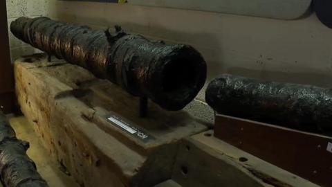 An old cannon muzzle on a wooden plinch along with another cannon next to it and a glimpse of another cannon on the ground.