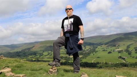 A man with tattoos, sunglasses and a plaited beard, with one foot on a stone, and the Peak District hills behind him.