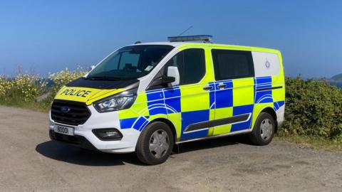 A Guernsey Police van parked next to a hedge along the island's coast on a sunny day. The van is yellow, blue and white. It has a licence plate of 80001.