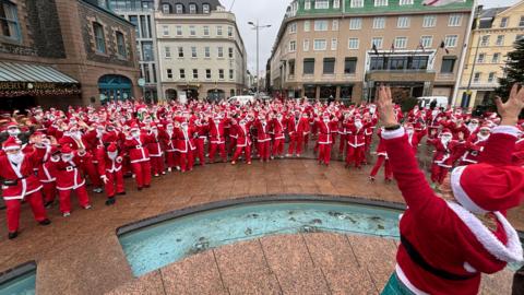 A crowd of people all dressed as Santa Claus standing outside with buildings surrounding them.