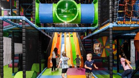 A picture of children playing in a brightly coloured soft play. There is a slide in the background and a tube for crawling through. 