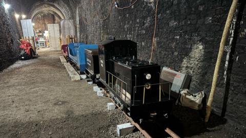A miniature railway on small tracks in an arch-shaped tunnel with dark brick walls. The front of the train is back with silver railings round the side.