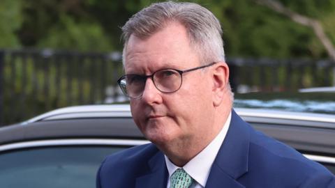 A man with short greying hair, the recededing hair line parted to the side, is getting out of a car. He is wearing black glass, a blue suit jacket, white tie and light green tie with a pattern. The top of the car behind him is black. In the background is a metal fence and some trees