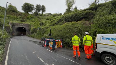 Workers in orange and yellow hi-viz stand next to a van at the entrance to Beaminster road tunnel. Overgrown embankments rise up either side of the two-way road. On the right is a small coned-off area with blue plastic barriers.