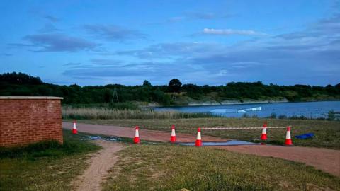 A lake with greenery around it, and orange traffic cones and a cordon in place near overflowing water.