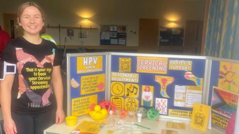 Cara Lawrence smiles at the camera while standing next to her cervical screening stand following a Parkrun. She has blonde hair which has been tied up and is wearing a running top that encourages people to attend their cervical screenings. She is wearing black leggings as well as an arm phone holder.