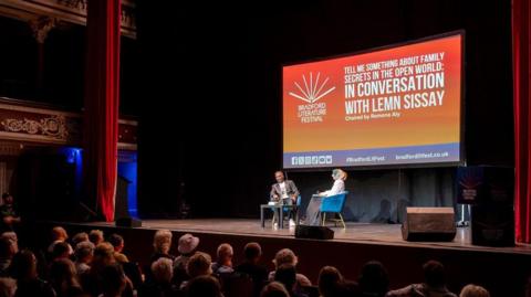 Two people on a stage in front of an orange whiteboard projected with "bradford literature festival" written in white and "in conversation with lemn sissay"