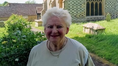 Ewelme historian Carol Sawbridge smiling for a picture in front of Ewelme Church. It is a sunny day.