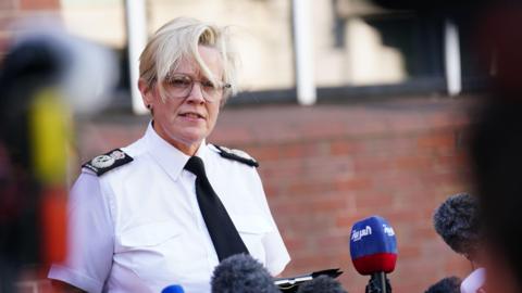 A woman with glasses and short blonde hair, wearing a black tie and policing uniform.