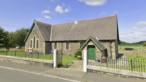 Lindisfarne Methodist Church in Christon Bank is an old stone building on one floor. It has six windows with arched tops, a single grey wooden door and green wooden double doors. It is surrounded by grass. There are two brown planters and two plant pots. There is a small tree near the double door and larger trees in the background. There are fields and trees further in the background. There is a white car in the distance. The church has a stone wall with metal railings and metal gates. There is a pavement in front.