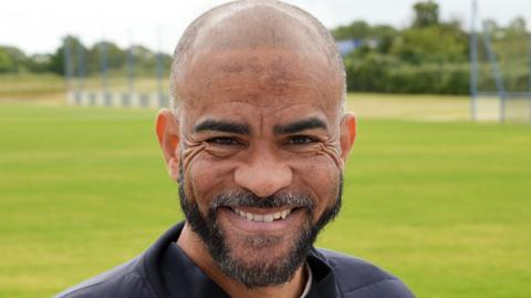 Kieron Dyer has a shaved head and a beard. He is wearing a navy jacket over a cream T-shirt and is smiling. He is standing in front of a football pitch at Southend United's training ground.