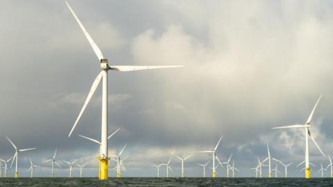 Large three-bladed white turbines out at sea. The water is dark and there are thick, dark clouds in the sky above.