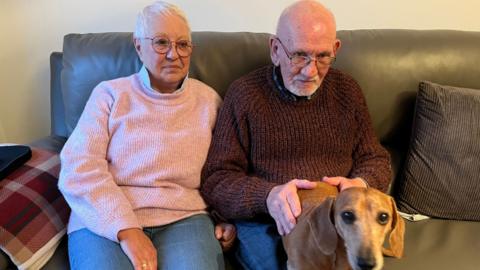 Mr and Mrs Clarke sitting on a dark brown sofa with Dachshund cross, Disney, sat on Mr Clarke's lap