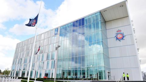 A blue flag with the Greater Manchester Police logo is flying at half mast outside a glass-fronted building which is the headquarters for the force. Two police officers are standing in front of the entrance. 