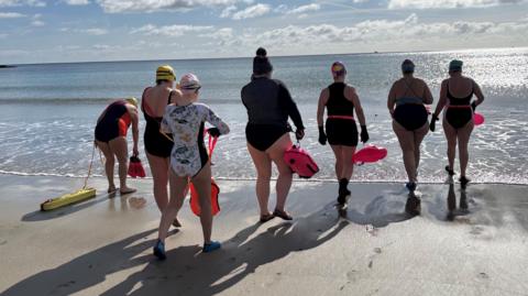 A group of women wearing swimming costumes and holding floats, walking into the sea on the beach