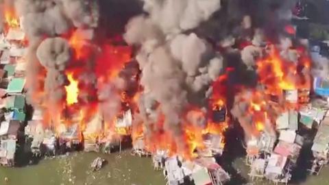 Smoke billowing as flames engulfing closely built houses next to water