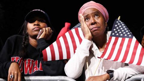 Two women stand with a hand against their head - one holds two US flags and wears a white hoodie, while the other wears a black university sweater reading Howard and has a black baseball cap