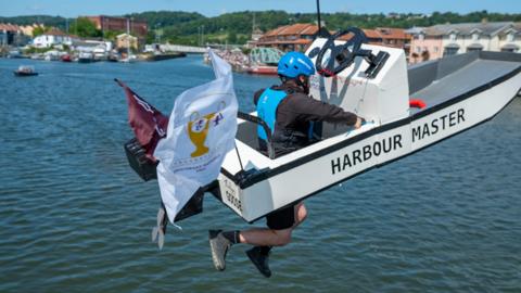 A man wearing a costume in the shape of a small speedboat that says Harbour Master on the side. He is in mid jump falling towards the water, and his legs are sticking out beneath the boat. He has a blue helmet and lifejacket on.