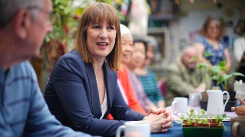 Rachel Reeves sat at a table surrounded by members of the public, with a plant pot in front of her and various cups and jugs.