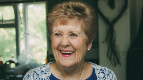 Beryl Howard wearing a blue top underneath a white lace cardigan, a silver necklace and bright pink lipstick. She is looking off to the left of the camera and laughing. She has short light brown hair and blue eyes.