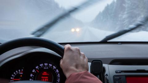 A hand on a steering wheel, wind screen visible although very foggy with wipers in motion, car headlights barely visible in distance