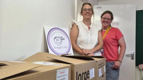 Two women standing next to cardboard boxes stacked against a wall