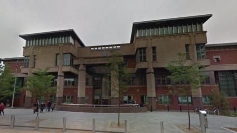 Exterior of modern red-brick and brown court building, with columns on its facade. A group of people is standing in front of the building to one side, while two trees stand in the foreground.