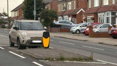 A car is parked on the chevrons in the middle of Luton Road in Dunstable with terraced houses on either side.