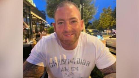 A picture of a man smiling at the camera. He has short black hair and a short brown beard. He is wearing a white T-shirt.