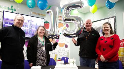 A group of people stand in a room with big silver number 15 balloons. Other balloons are on the ceiling. A cake with candles is in the middle of the table.