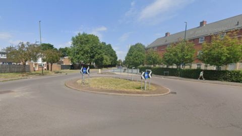Streetview image of a roundabout with black and white chervron signs and blue and white one-way arrows. On the left are 1960s social housing units, and on the right, more modern, three-storey flats with a hedge in front. 