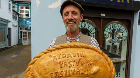 A person wearing a flat cap and a chequered shirt holding a large pasty which says 'Redruth Pasty Festival!' written on it in pastry.