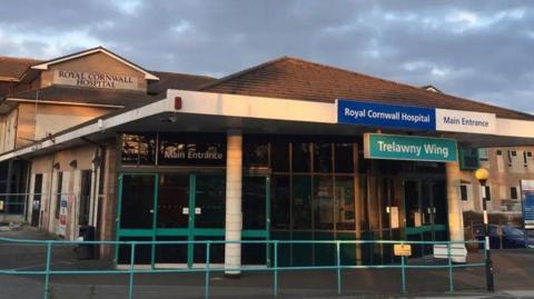 The front of the main entrance of the Royal Cornwall Hospital. There are glass doors which say main entrance on them. There is a blue sign hanging down from the roof which says 'Trelawny Wing'.  