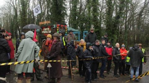 A group of campaigners stood in a field, with a line of trees behind them. They are stood behind yellow and black tape.