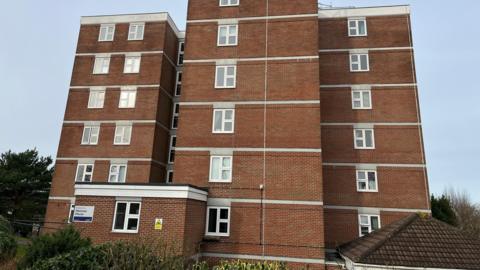 A six-storey block of flats. The building is made of brown brick with uniform windows in line going up its three main columns. 