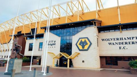 Exterior of Molineux Stadium showing black letting on the building saying 'Wolverhampton Wanderers F.C', and 'Billy Wright stand', there is also the club badge - a black wolf head with orange background in an hexagonal shape.