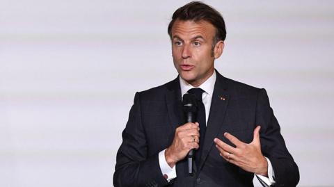 France's President Emmanuel Macron in a white shirt and suit holds a microphone and talks to an audience