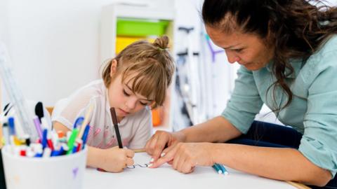 A teacher leaning over and helping a child who is holding a pen and drawing on some paper in a classroom setting.