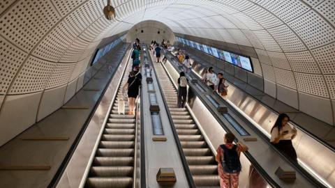 General view of a triple escalator at Liverpool Street station. Two are going up and one is going down. Various passengers are standing on all escalators.