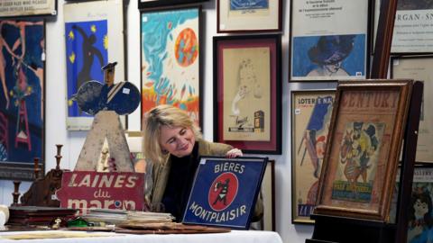 A woman sits at a table holding and looking at a enamel sign. All around her are prints and signs from the 20th century.