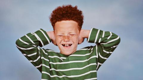 Boy wearing a striped green and white top and red hair covers his ears with his hands