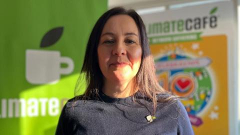 Jess Pepper, a dark-haired woman in a black sweater and a Climate Café pin badge, stands in front of a the café signage which depicts a coffee mug and a colourful 10-year anniversary graphic.