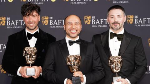 Three men stand on the 2026 BAFTA red carpet in black tuxedos. Each of the three hold a BAFTA award in the shape of a gold theatrical mask.