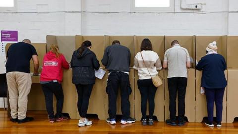 Shown from behind, a line of people face cardboard voting booths
