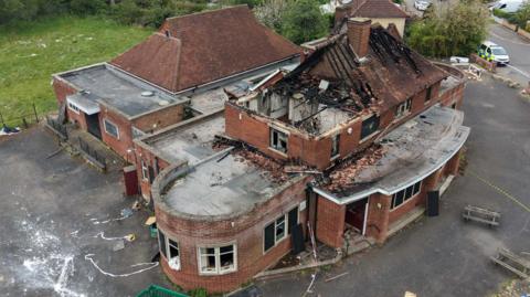 Drone footage of a brick built building, with curved walls, showing a destroyed roof. A police car is to the right and other buildings down a street. 