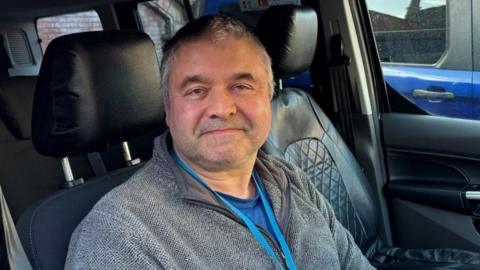 There is a man sat in a taxi with black leather interior seating. He has short grey hair, a grey stubbly beard, and a grey fleece jacket. He also is wearing a blue t-shirt and a blue lanyard string.