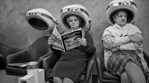 A black and white image of two women sitting in the hairdressers with heating cones on their heads. They are older women, one is reading a copy of Northern Life magazine and both have a rather cross expression on their faces 